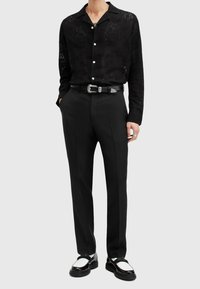 Camicia nera a maniche lunghe con motivo floreale, bottoni bianchi e colletto a punta, abbinata a pantaloni neri e scarpe bicolori in contrasto, nere e bianche.