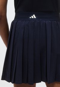 Marineblauwe geplooide rok met een elastische tailleband met het Adidas-logo in wit. Glad materiaal met een gelaagde textuur.