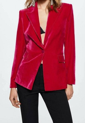 Frau trägt einen glänzenden roten Samtblazer über einem schwarzen Top und schwarzen Hosen, accessoriert mit langen hängenden Ohrringen.