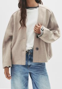 Femme portant un manteau beige boutonné par-dessus un haut blanc et une chemise à manches longues rayée, accompagnés d'un jean en denim bleu.