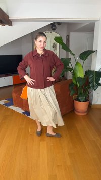 Femme debout dans le salon, les mains sur les hanches, portant une veste bordeaux, une jupe beige et des ballerines grises, près d'une grande plante en pot et d'un canapé orange.