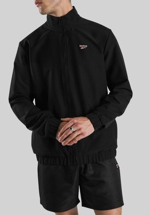Mann in schwarzer Reebok-Zip-Up-Jacke und passender Shorts, die Hände vor dem Körper verschränkt, trägt einen silbernen Ring am linken kleinen Finger.