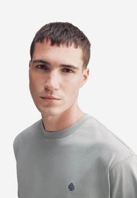 T-shirt grigio chiaro a girocollo con maniche corte, caratterizzato da un logo ad albero discreto sul petto e una texture in cotone morbido.