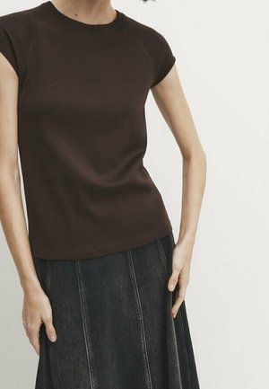 Femme portant un haut marron foncé ajusté à manches courtes et une jupe en denim foncé taille haute avec des coutures verticales, debout devant un fond uni.