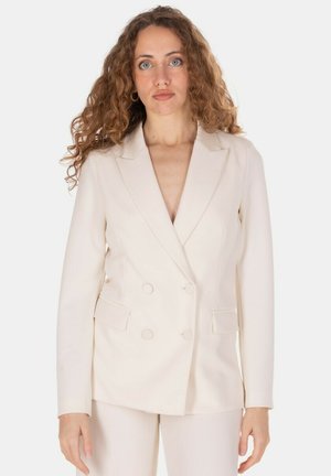 Donna con capelli ricci che indossa un blazer crema doppiopetto e pantaloni abbinati, in piedi davanti a uno sfondo bianco semplice.
