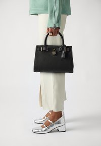 Sort tekstureret læder håndtaske med to håndtag, guld hardware og et dekorativt mærke. Sammen med sølvmetalliske sko og cremede bukser.