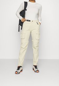 Ljusbeige cargobyxor med flera fickor, ett bälte och en slim fit. Stylistiskt kombinerade med en vit långärmad topp och flerfärgade sneakers.