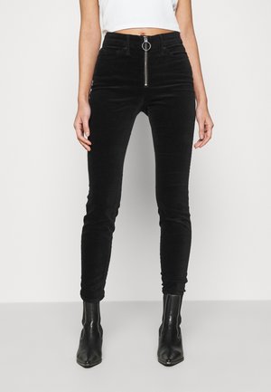 Femme portant un pantalon noir skinny taille haute avec fermeture éclair avant et bottines noires, debout devant un fond clair uni.