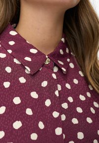 Femme portant une chemise col marin maroon à pois blancs avec un petit bouton rond en or au col, avec de longs cheveux bruns visibles.