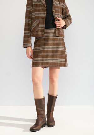 Femme portant une jupe à carreaux marron et un blazer assorti avec un haut noir, associée à des bottes en cuir marron mi-mollet, debout sur un fond blanc.