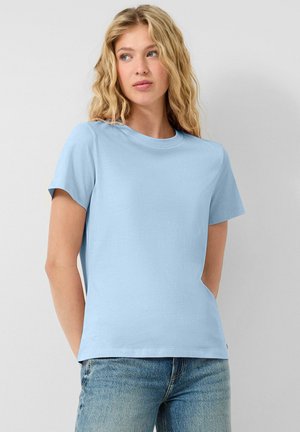 Jeune femme aux cheveux blonds ondulés portant un t-shirt à manches courtes bleu clair et un jean bleu, regardant légèrement de côté.