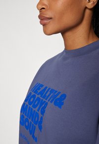 Mörkblå sweatshirt med korta ärmar, rund halsringning, med en djärv blå text: "HÄLSA & KROPP & SINNE & SJÄL." Mjuk, strukturerad tyg.