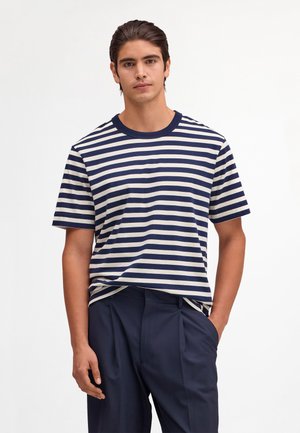 Giovane uomo che indossa una maglietta a righe orizzontali blu navy e bianche e pantaloni blu navy plissettati, in piedi con una mano in tasca su uno sfondo bianco.