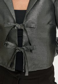 Metallisch zilveren cropped jack met twee voorkantstrikken en een gestructureerde afwerking, gedragen over een zwarte top en gecombineerd met donkere broek.