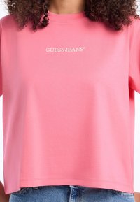 Růžové bavlněné tričko s výstřihem do kulata a krátkými rukávy, s vyšitým logem "GUESS JEANS®" na přední straně. Hladká textura, pohodlný střih.
