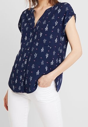 Femme portant un chemisier à manches courtes bleu marine avec de petits motifs de cactus blancs et de cœurs roses, assorti à un pantalon blanc.