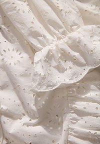 Witte stof met ingewikkelde oogjesborduurwerk met florale patronen, gerimpelde details en een gegolfde rand op het geplooide gedeelte.