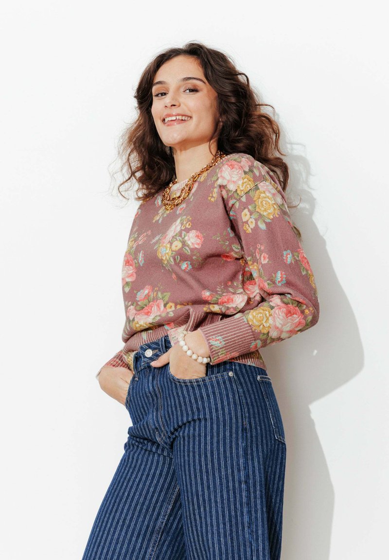 Femme souriante aux cheveux bouclés portant un pull à fleurs, un jean rayé, un collier en chaîne dorée et un bracelet de perles, posant les mains dans les poches.