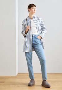 Camicia button-up a righe blu chiaro e bianco, t-shirt bianca, jeans blu chiaro e scarpe grigie. Modello in un ambiente minimalista.