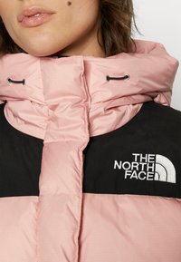Veste doudoune rose avec une partie supérieure noire portant le logo "The North Face". Le tissu a une texture lisse et comprend des cordons de serrage ajustables.