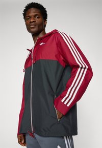 Rød og svart lett jakke med glidelås, justerbar hette og to sidelommer. Har tre hvite striper på ermene.