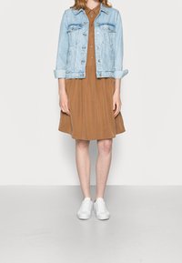 Světlá modrá džínová bunda přes hnědé šaty. Šaty mají knoflíkový přední díl a outfit je doplněn bílými teniskami.