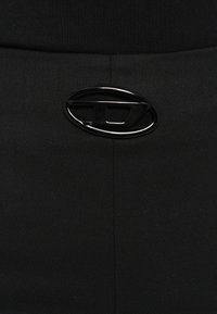 Melns audums ar faktūru apdari, pie jostas līnijas redzams spīdīgs melns ovāls logotips kā ievērojama detaļa.