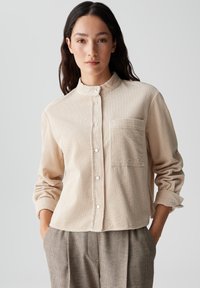 Crème corduroy overhemd met een staande kraag, knoopsluiting aan de voorkant en een enkele borstzak. De stof heeft een geribde textuur en ontspannen pasvorm mouwen.