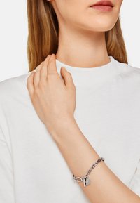 Silverfärgad kedjebracelet med rund berlock, som har en blandning av polerade och strukturerade länkar, bärs på en ljus hudton mot en vit topp.