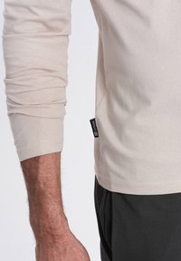 Helles beige Langarmshirt mit gerippten Bündchen; seitliches Etikett sichtbar. Weicher, dehnbarer Stoff; getragen mit dunkelgrauen Hosen.