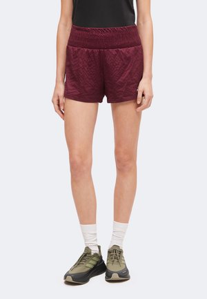 Persoon draagt bordeauxrode sportshort, witte sokken, olijfgroene en zwarte sneakers en een zwart shirt, staand tegen een lichte achtergrond.