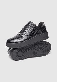 Las zapatillas negras con estampado de cocodrilo presentan un exterior de cuero suave y brillante, un diseño de cordones y una suela de goma texturizada con acentos circulares.