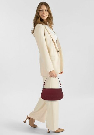 Femme portant un tailleur crème et des talons marron clair, tenant un sac à main bordeaux, souriante et marchant devant un fond clair uni.