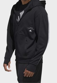 Svart zip-up hoodie i texturerat tyg, med framdragkedja, sidfickor och vita accentremsor längs ärmarna.