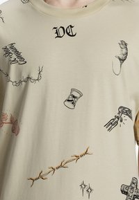Camiseta de algodón beige con estampados gráficos en negro y rojo, que incluyen un ratón, una rosa, un reloj de arena y varios diseños en líneas.