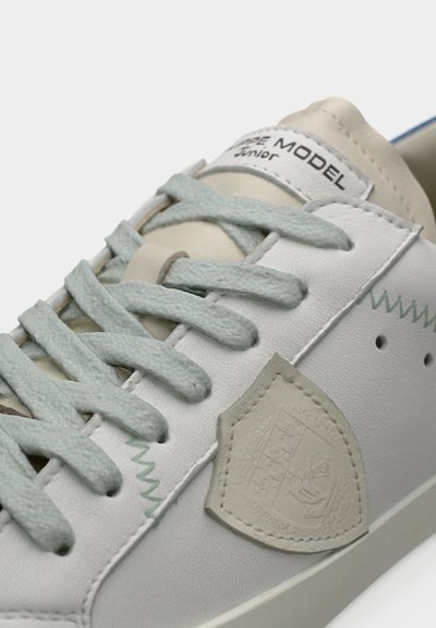 Λευκό δερμάτινο αθλητικό παπούτσι με γαλάζια κορδόνια, διακοσμητικές ραφές και υφασμένο μπεζ μέρος που μοιάζει με ασπίδα. Παρουσιάζει διακριτικές λεπτομέρειες σχεδίασης.