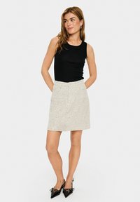 Top fără mâneci de culoare neagră, asortat cu o fustă texturată de un bej deschis cu un model pistruiat, completat de pantofi cu vârful ascuțit de culoare neagră.