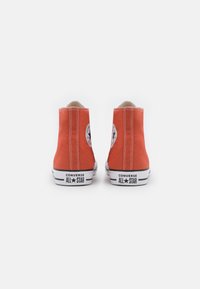 Sapatilhas de cano alto em laranja, em tela, com costura branca e solas de borracha. Apresenta um logo circular e o rótulo "CONVERSE ALL STAR" no calcanhar.