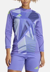 Paarse sporttop met lange mouwen, versierd met geometrische patronen in marineblauw en lichtgroen. Gekleed met bijpassende paarse shorts.