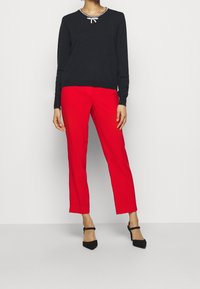 Pull marin bleu avec un nœud et des embellissements en strass, associé à un pantalon rouge vif et des chaussures à talons noirs, créant un ensemble contrasté.