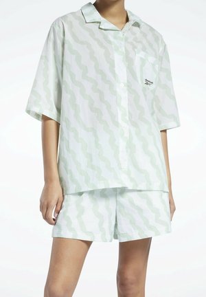 Osoba nosící bílou volnou košili s krátkým rukávem na knoflíky a odpovídající šortky se světle zeleným cikcak vzorem a logem Reebok na kapse.