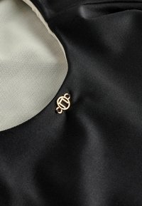 Crni satenski materijal s glatkom teksturom ima umetak od mrežaste tkanine i zlatne metalne detalje. Istaknuta kružna dizajnerska nijansa na zatvaranju.