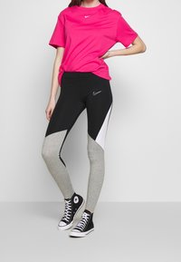 Růžové tričko s krátkým rukávem značky Nike, doplněné černými a šedými legínami s bílým vzorem a logem, spolu s černými vysokými teniskami.