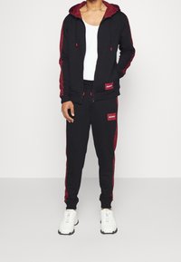 Hombre vistiendo un chándal negro con rayas rojas en el patrón, camiseta sin mangas blanca, zapatillas blancas, de pie contra un fondo claro y liso.