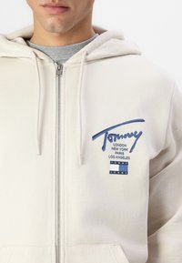 Kremast hoody s patent zatvaračem, kapuljačom s povlačenjem i plavim logom koji sadrži imena gradova i pravokutnu oznaku. Glatka tekstura i ležeran dizajn.