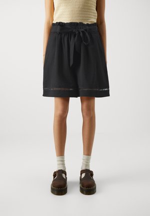 Kvinde iført sorte knælange shorts med bælte, beige striktrøje, hvide sokker og brune sko med spænde, stående mod en ensfarvet baggrund.