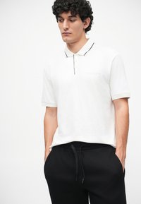 Biele polo tričko s čiernymi detailami a logom, s krátkymi rukávmi, v kombinácii s čiernymi teplákmi s viazacím pásom. Minimalistický dizajn.