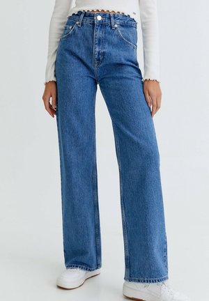 Woman wearing high-waist wide-leg blue jeans, white long-sleeve cropped top, and white sneakers, standing against plain background.