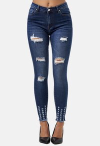 Jeans ajustados de mezclilla azul oscura, con áreas desgastadas y dobladillos deshilachados, además de detalles decorativos. Terminado con un cierre de botón.