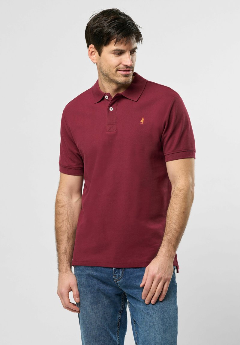 Burgundi polo marškiniai su apykakle ir trumpomis rankovėmis. Yra mažas siuvinėtas logo ir tekstūruotas audinys. Dėvimi su mėlynais džinsais.